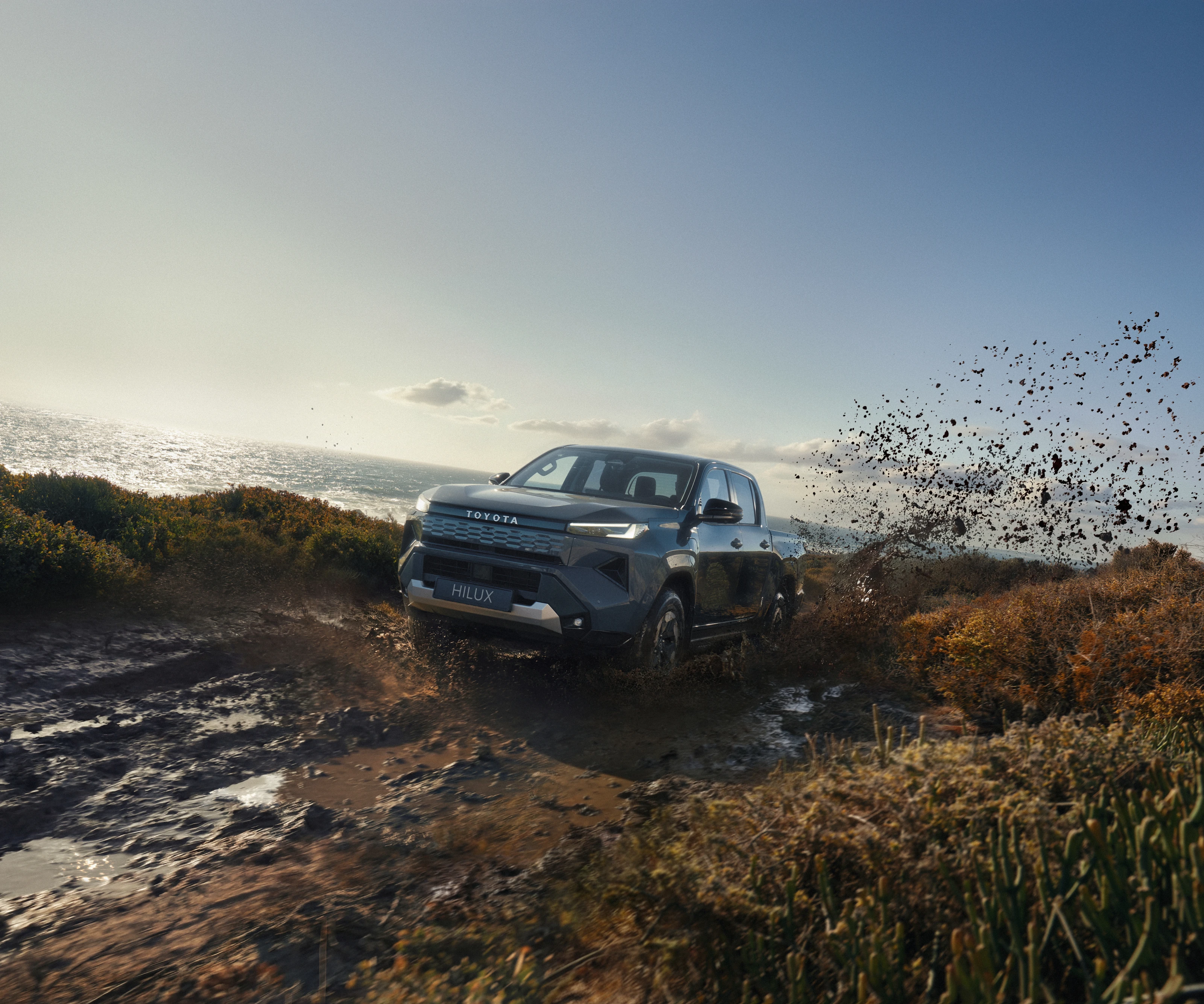 Grey Toyota Hilux BEV throwing mud on a coastal track
